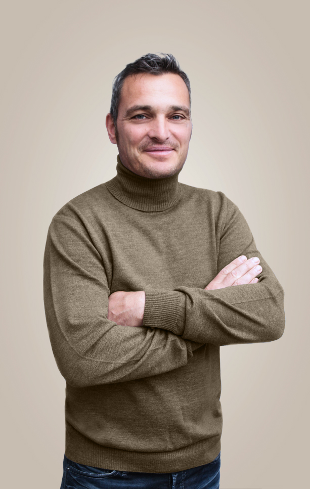 Mann in camelfarbenem Pullover hat die Arme verschränkt und lächelt.